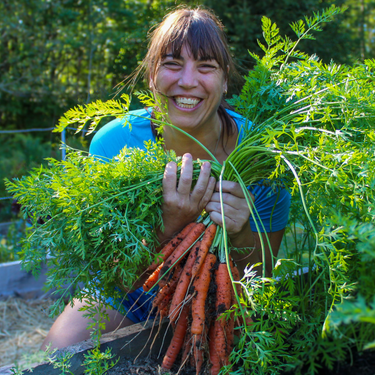 A smiling woman in a blue shirt holds a freshly harvested bunch of orange carrots with green leafy tops in a garden setting.