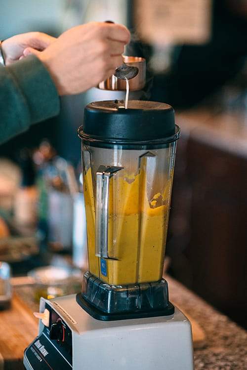 hand pouring milk into blender with pumpkin puree using spoon and measuring cupt