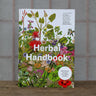 Herbal Handbook with recipes, tinctures, and balms with bright illustrated cover on a wooden table.